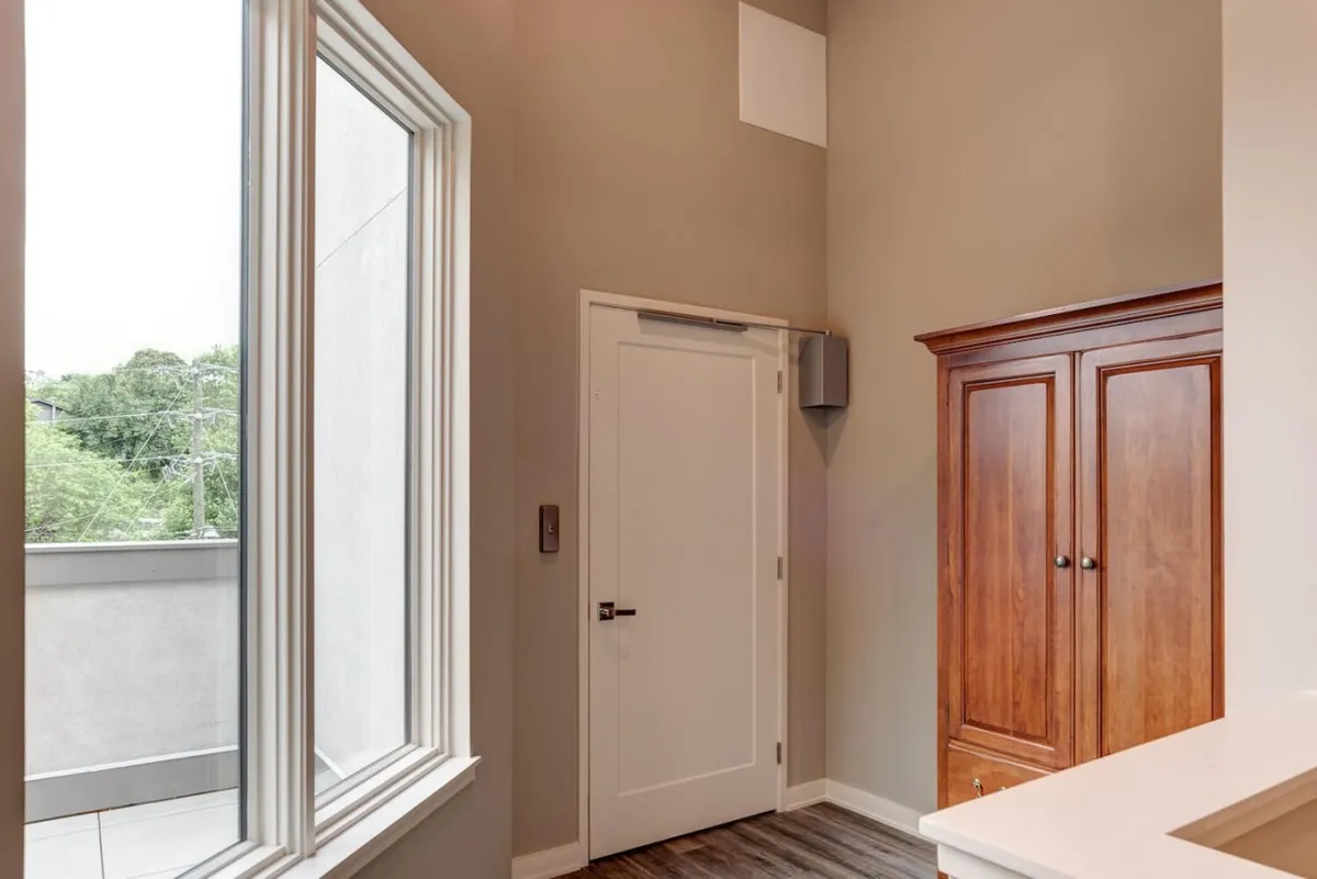 This image features a large window offering a view of the outdoors, a wooden cabinet, and a white door, all arranged to create a welcoming entryway or transition space. The calm, serene mood is enhanced by natural light streaming in, suggesting a peaceful environment both inside and outside the property.