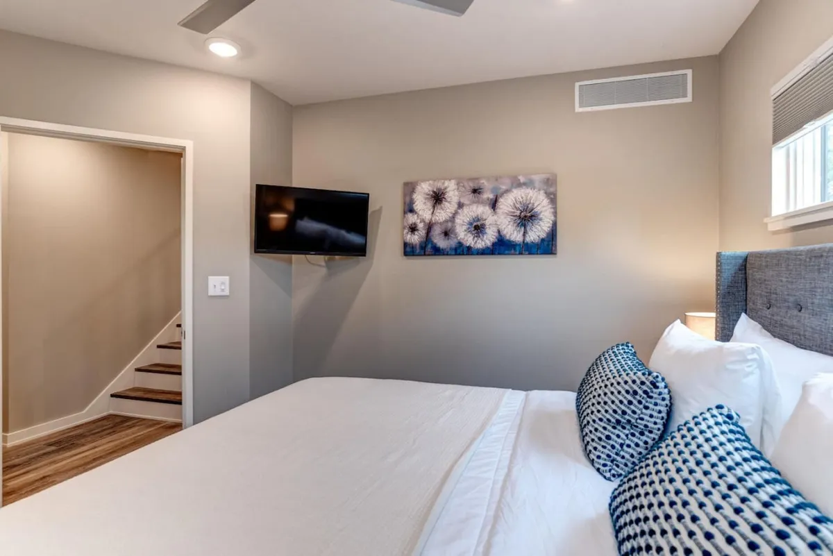 A bedroom scene with a well-made bed, decorative pillows, a TV, wall art, and a visible staircase, suggesting multi-level living. The inviting setup reflects a space ideal for unwinding after exploring the city, with architectural cues like the staircase hinting at easy access to upper levels or a rooftop.