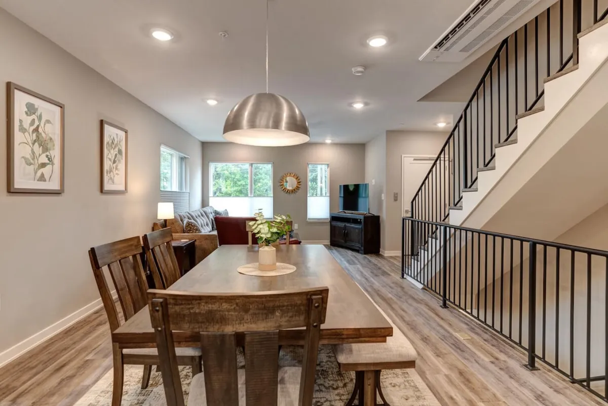 A cozy living area featuring a large dining table, comfortable sofa, television, and wall art. The room’s layout is ideal for both casual group relaxation and formal meals. The staircase in the background hints at the home’s vertical, multi-level structure.