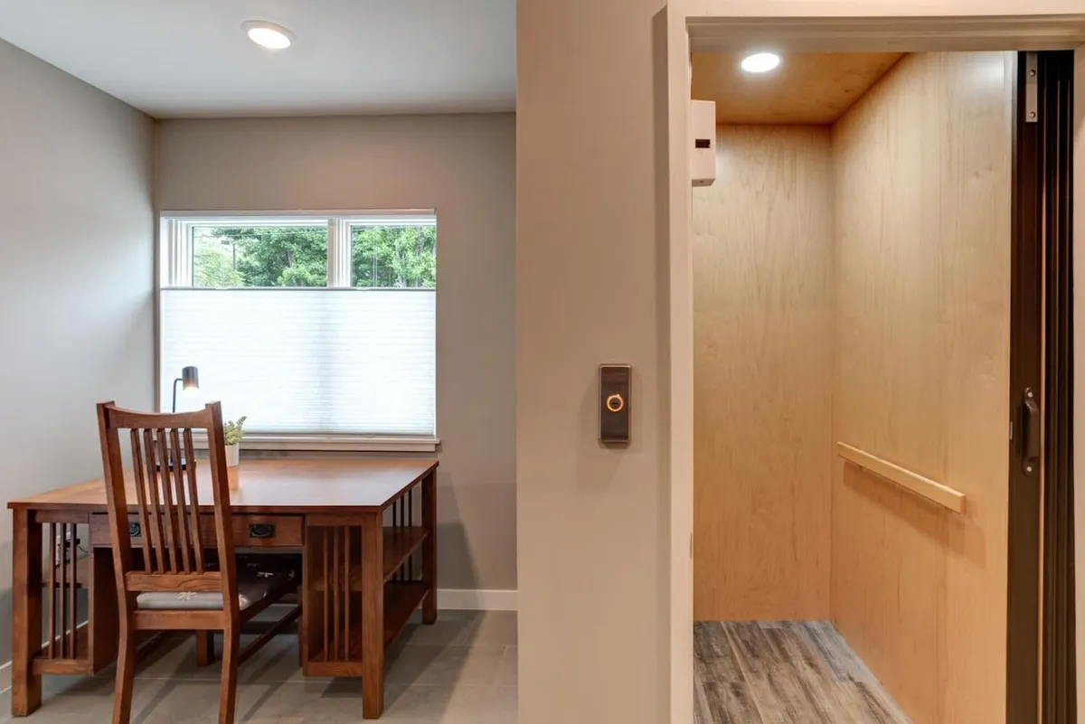 A calm, productive interior corner featuring a wooden desk set near a window and, most importantly, a clear view of the elevator entrance. The mood is serene, emphasizing ease of movement and accessibility within the home.