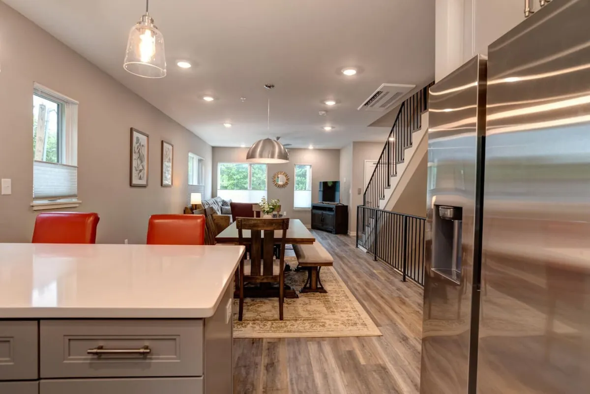 This image features a spacious kitchen island and dining table, both central gathering spots for groups. The open layout is accented by stylish light fixtures and a visible staircase, emphasizing the multi-level nature of the townhome. The scene conveys an inviting and warm atmosphere designed for entertaining, shared meals, or impromptu celebrations.