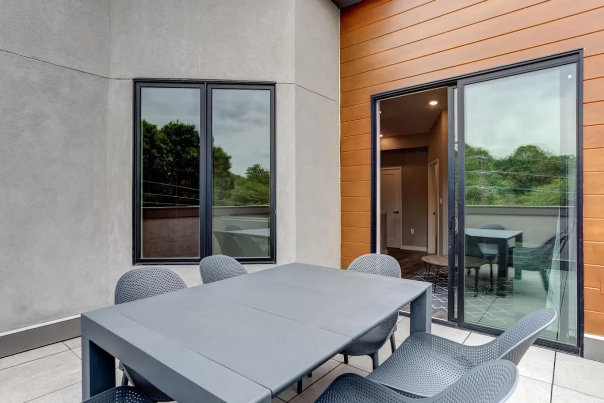 Outdoor dining table and chairs set directly beside large glass doors that open into the interior, with greenery surrounding the patio. This image highlights the seamless transition between indoor comfort and outdoor living, emphasizing versatility and connection with nature.