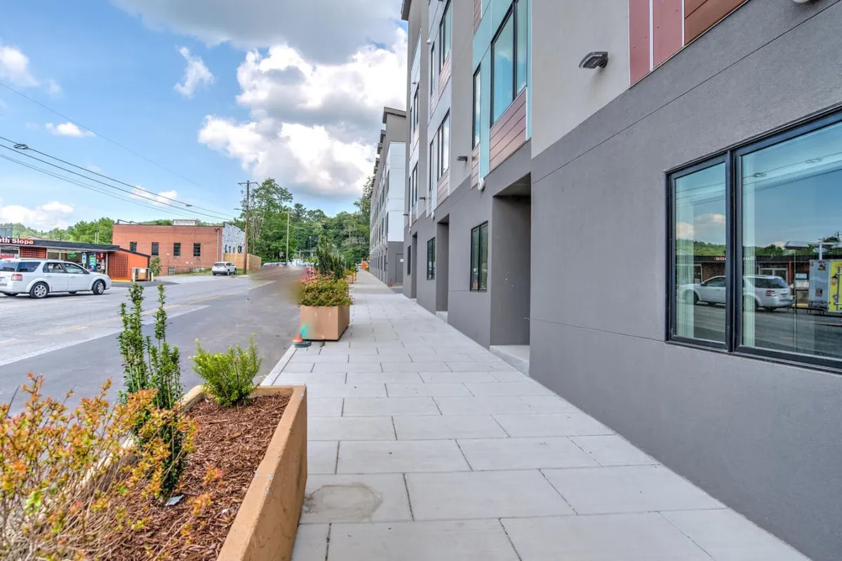 This image captures a modern building with striking architectural features, situated along a lively sidewalk adorned with planters. The street view is bustling and inviting, suggesting a vibrant and walkable neighborhood.