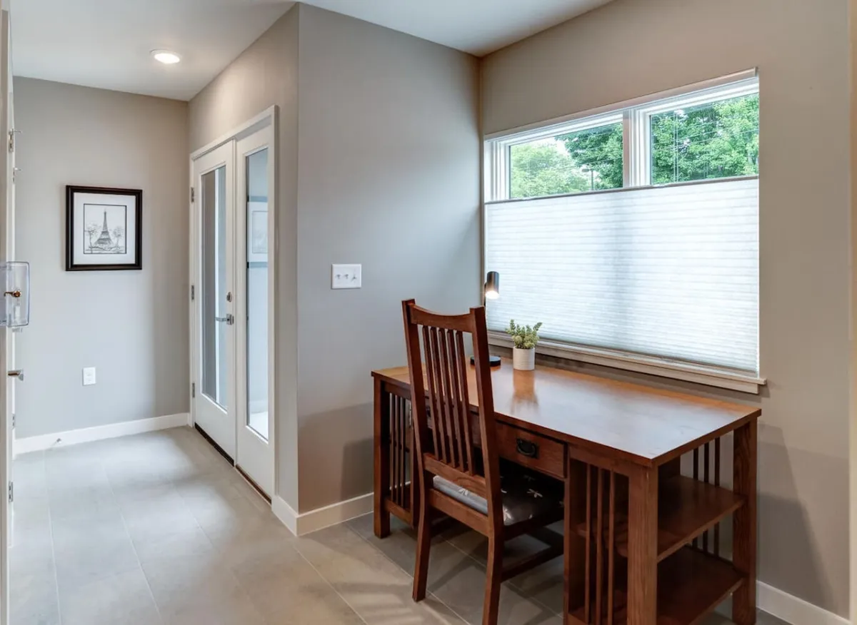 A bright sitting area featuring a wooden desk and chair, situated next to a large window that lets in natural light. Framed artwork decorates the wall, creating an inviting and serene indoor lounge perfect for reading, working, or relaxing while enjoying city views.