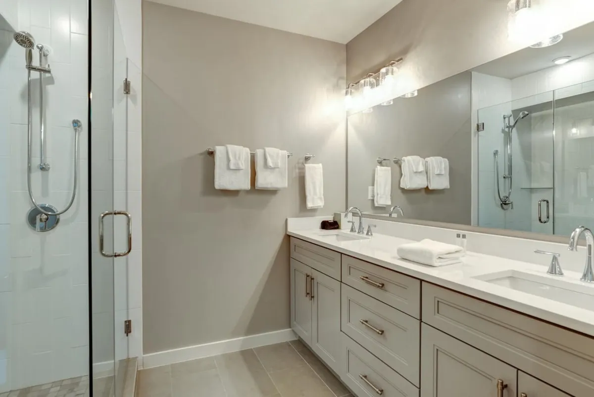 A spacious bathroom with a double sink vanity, expansive mirror, glass shower enclosure, and plush towel rack. The room feels luxurious and tranquil—perfect for relaxing self-care routines.