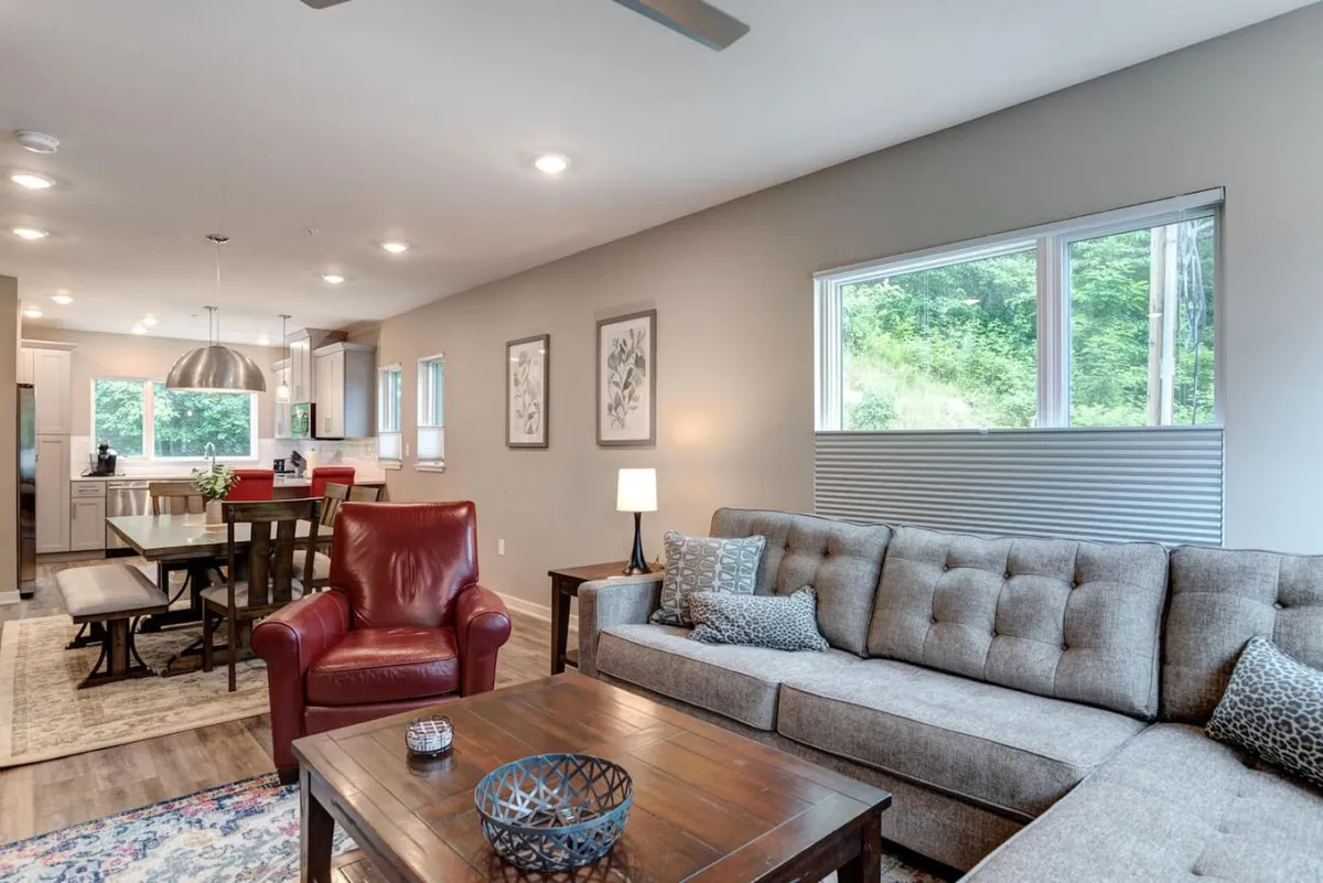 A spacious living and dining area featuring a comfortable sofa, red armchair, dining table, and a kitchen visible in the background. Large windows allow daylight to flood the space, while stylish artwork and furnishings set a sophisticated yet cozy backdrop for socializing or unwinding after an urban adventure.