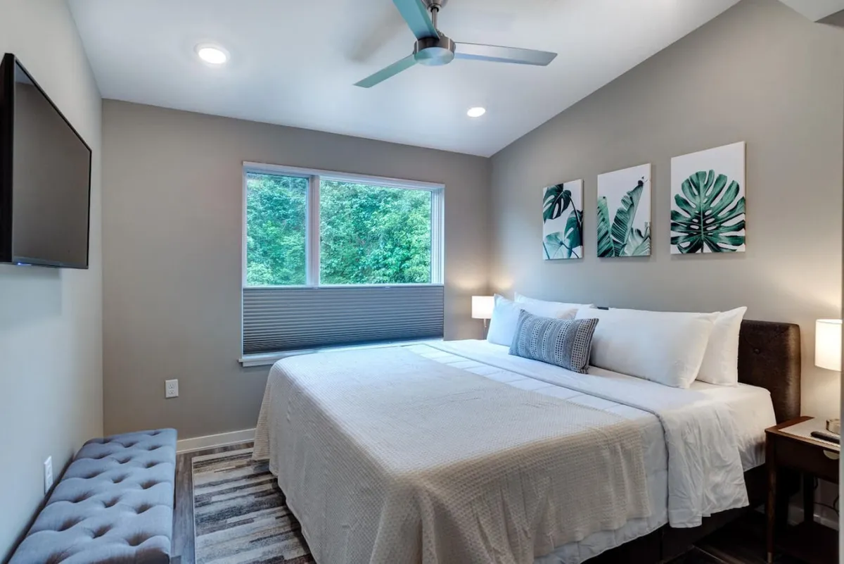 A serene bedroom with a comfortable bed, textured bedding, large window with city views, wall art, nightstands, and a television. The decor is elegant yet calming, with plenty of natural light enhancing the peaceful vibe.