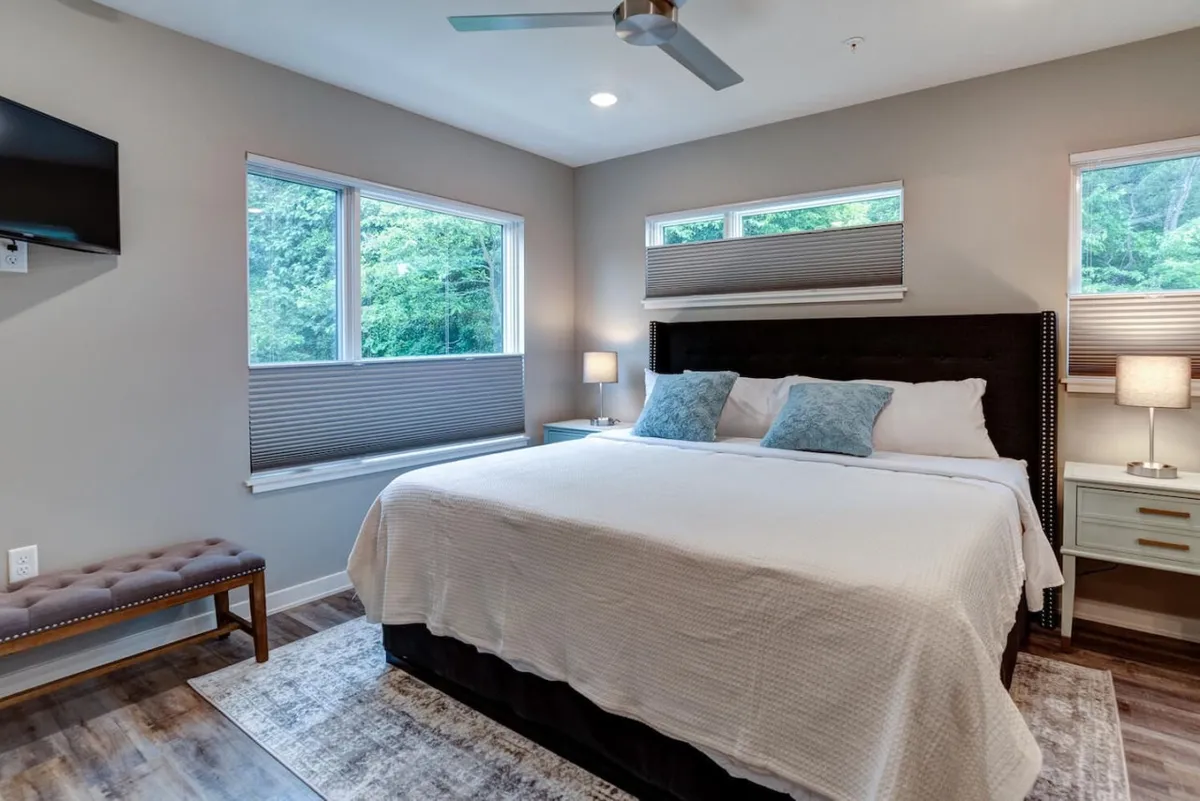 A tranquil king-sized bedroom featuring plush white bedding with blue accent pillows, a wooden bench at the foot of the bed, a nightstand with a lamp, and a wall-mounted television. Ample natural light streams in through the windows, highlighting the restful decor.