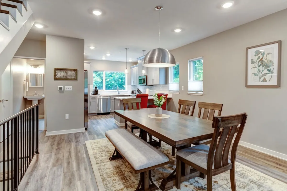 This image presents a dining table set in a welcoming, open area that flows seamlessly into a contemporary kitchen. The kitchen countertop and large windows—with views of lush greenery—emphasize natural light and an inviting atmosphere ideal for family meals and entertaining guests. Sophisticated furnishings and thoughtful design details add to the chic ambiance.